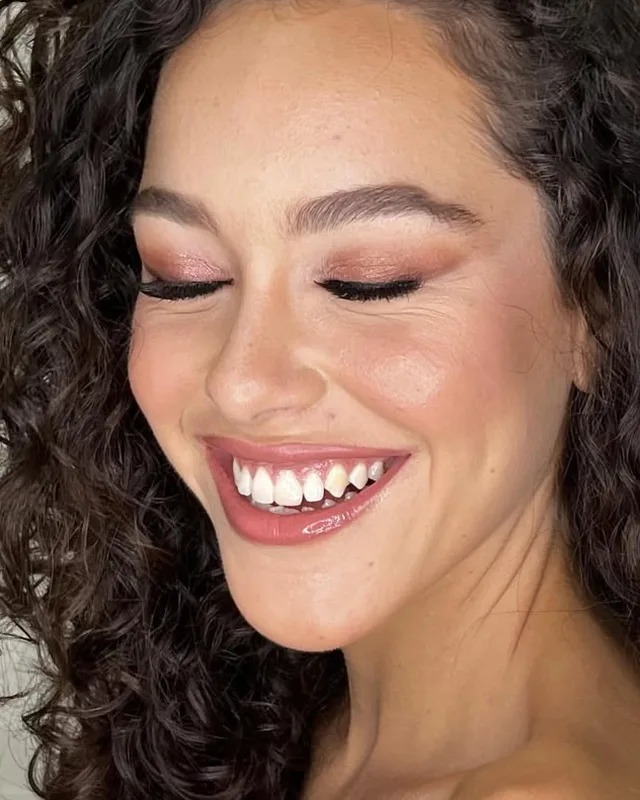 Cliente de cabelo cacheado sorrindo, de olhos fechados, com maquiagem iluminada
